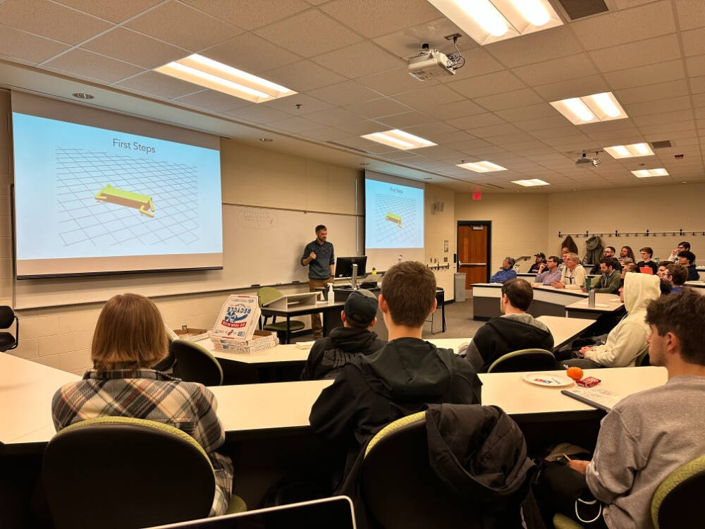 A university classroom features a lecturer standing at the front, speaking to a group of attentive students seated at curved rows of desks. Two large projection screens display a slide titled "First Steps" with a yellow 3D object on a grid.